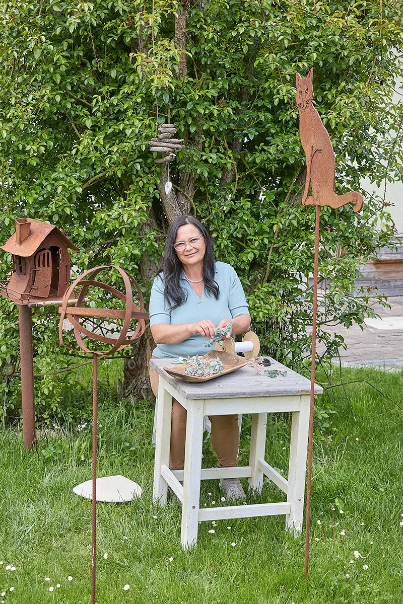 Frau bei Handarbeit am Tisch im Garten, umgeben von Metallkunstwerken und Vogelhaus.