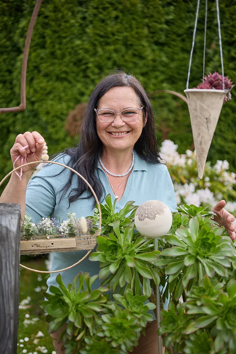 Ältere Frau mit Brille hält Dekoobjekt im Garten, lächelt in die Kamera.