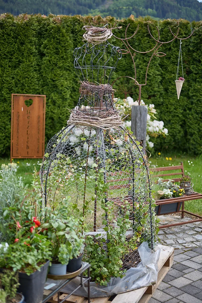 Ein Drahtkleid als Gartendeko mit Pflanzen, umrahmt von Gruen und Gartenrequisiten.