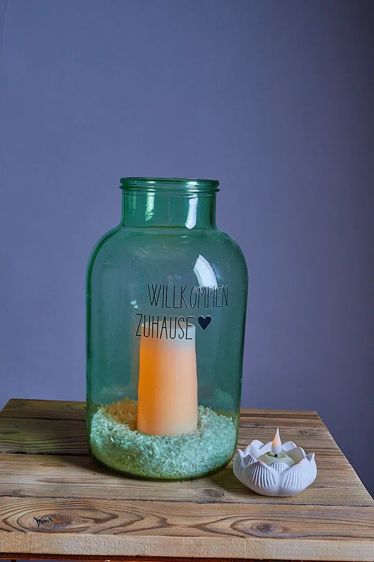 Grünes Glas mit Kerze und "Willkommen Zuhause"-Text, auf Holztisch mit kleinem Teelichthalter.
