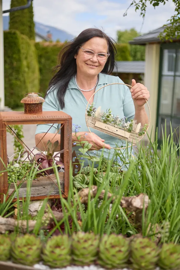 Frau im Garten mit Pflanzen und Dekorationen, sanft lächelnd. – handgearbeitete Deko Trieben